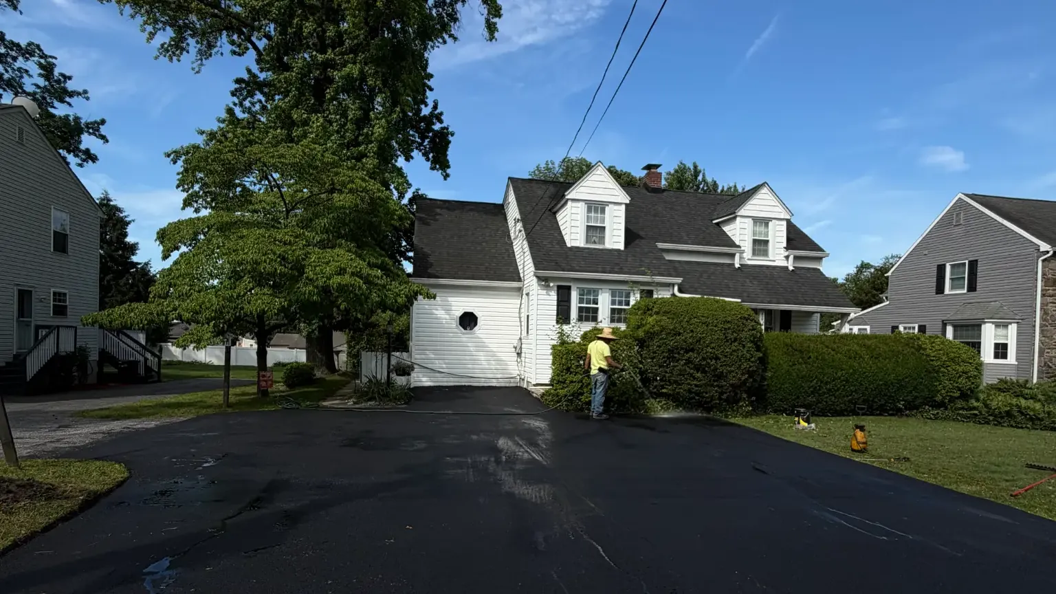 Freshly sealcoated residential driveway in front of a white two-story home, with a crew member finishing the edge by hand